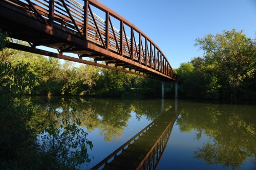 A metal bridge arches over a calm river, reflecting the blue sky and surrounding greenery.