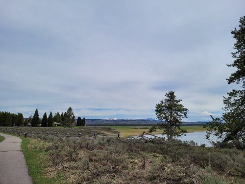 A scenic view of a pathway alongside a river, with trees and mountains in the background under a cloudy sky.