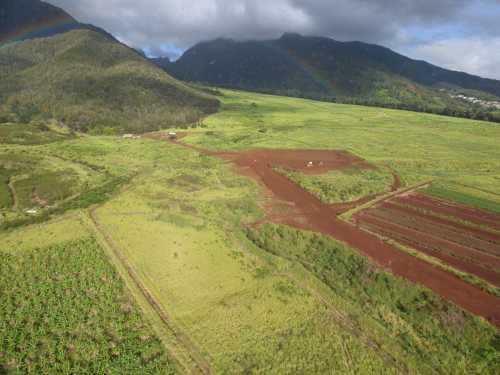 Aerial view of lush green fields and red soil, with mountains in the background and a rainbow in the sky.