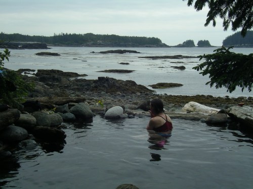 A person relaxes in a natural hot spring by the ocean, surrounded by rocks and trees under a cloudy sky.