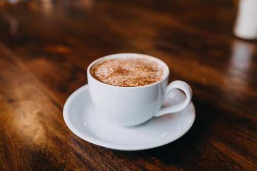 A white cup of coffee topped with cinnamon sits on a saucer on a wooden table.