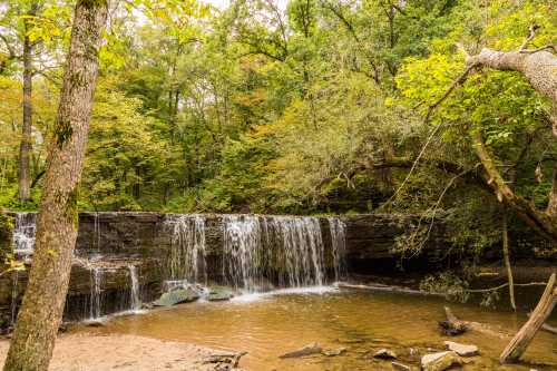 A serene waterfall cascades over rocks into a calm pool, surrounded by lush green trees and autumn foliage.