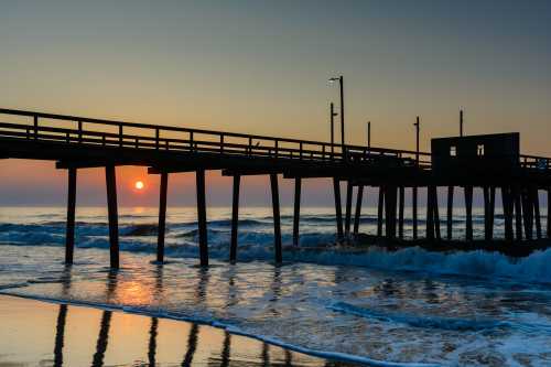 A wooden pier extends over calm waters at sunset, with gentle waves lapping at the shore and a warm glow in the sky.