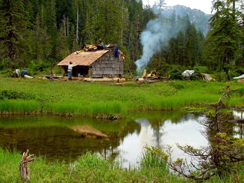 A rustic cabin under construction near a serene pond, surrounded by lush greenery and distant mountains. Smoke rises from a fire.