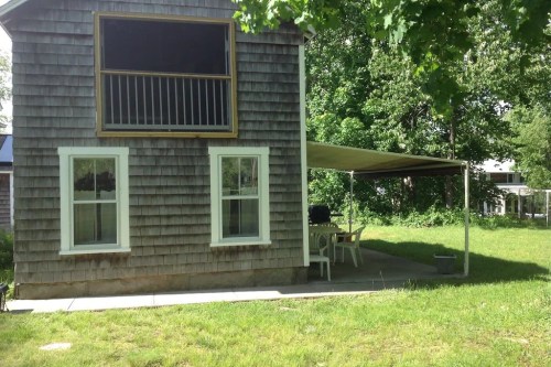 A two-story wooden house with a porch, surrounded by greenery and a shaded outdoor seating area.