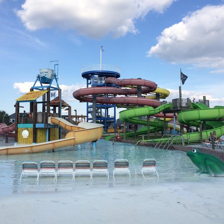 A water park featuring colorful slides, a shallow pool, and lounge chairs under a blue sky with fluffy clouds.