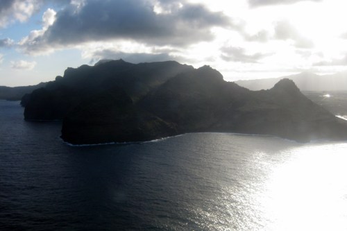 A scenic view of a rugged coastline with mountains, reflecting sunlight on the water under a cloudy sky.