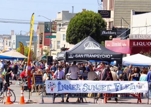 A bustling street fair with a large banner reading "Inner Sunset Flea" and crowds enjoying various stalls and activities.