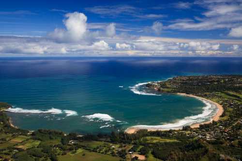 Aerial view of a serene coastline with waves, sandy beach, and lush green hills under a blue sky with clouds.