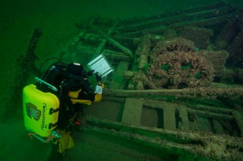 A diver examines a shipwreck underwater, surrounded by marine growth and debris.