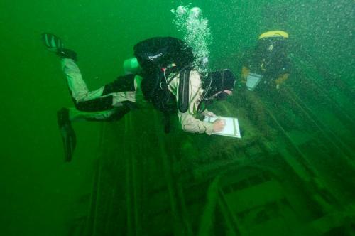 A diver writes notes underwater, surrounded by green water and bubbles, with another diver in the background.