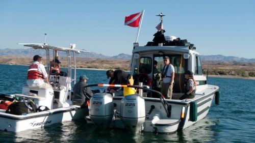 Two boats on calm water, one with divers preparing gear and the other with people in life jackets. Mountains in the background.