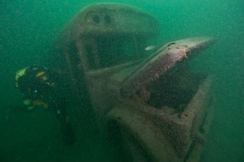 A diver explores a submerged, rusted vehicle in murky water, surrounded by bubbles and sediment.