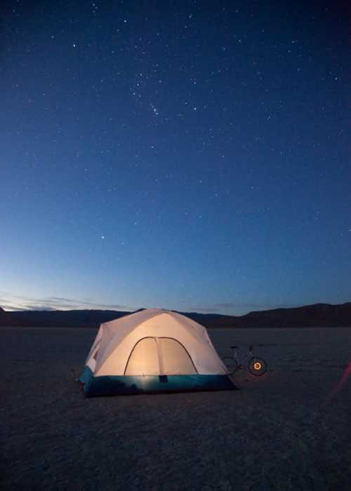 A tent illuminated at night under a starry sky, with a bicycle nearby on a barren landscape.