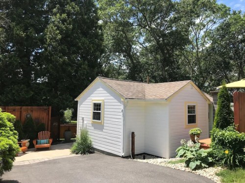 A small white shed with a brown roof, surrounded by greenery and a stone pathway in a sunny outdoor setting.