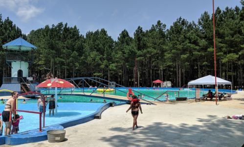 A sunny outdoor pool area with people swimming, water slides, and shaded seating surrounded by trees.