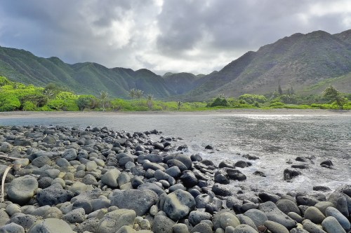 A rocky shoreline with smooth stones, leading to a calm water body surrounded by lush green mountains under a cloudy sky.