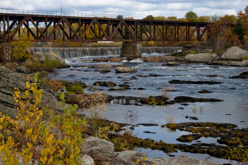 A rusted train bridge spans a river with waterfalls, surrounded by autumn foliage and rocky shores.