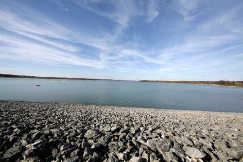 A calm lake with a rocky shoreline under a blue sky with wispy clouds.