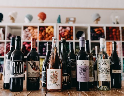 A collection of wine bottles arranged on a wooden table, with colorful yarn displayed in the background.