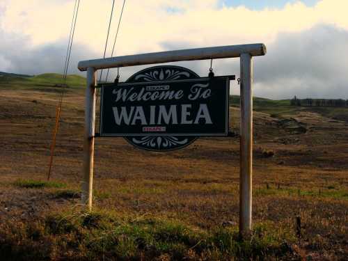 Sign welcoming visitors to Waimea, set against a grassy landscape and cloudy sky.