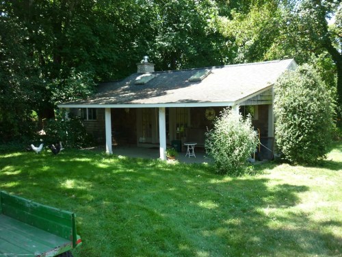 A small, rustic house surrounded by trees and greenery, with a porch and a well-maintained lawn.