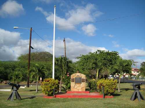 A memorial with a plaque surrounded by greenery, flagpole, and cannons under a blue sky with clouds.