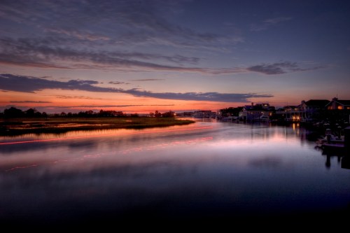 Serene sunset over a calm waterway, reflecting vibrant colors and surrounded by lush greenery and homes.