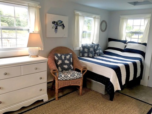 Cozy bedroom with a striped bed, wicker chair, white dresser, and natural light from windows.
