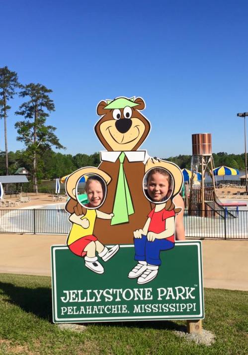 Two children pose in a cutout sign featuring a bear mascot at Jellystone Park in Pelahatchie, Mississippi.