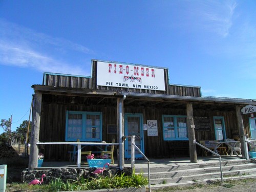 A rustic building with a sign reading "Pie Town, New Mexico," featuring blue windows and a porch with flamingo decorations.