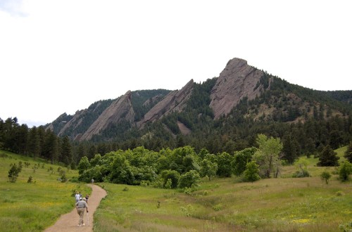 A hiker walks along a dirt path with lush greenery and rocky mountains in the background under a cloudy sky.