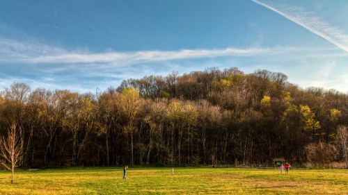 A grassy field with three people in the foreground and a wooded hill in the background under a clear blue sky.