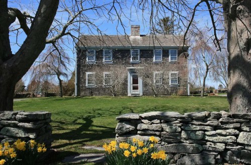 A historic wooden house surrounded by trees and blooming daffodils, with a stone wall in the foreground.