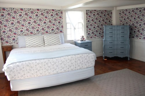 Bright bedroom with floral wallpaper, a light blue bedspread, and a vintage blue dresser. Natural light from a window.