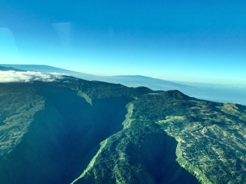 Aerial view of rugged mountain terrain under a clear blue sky, with distant peaks visible on the horizon.