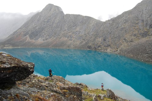 Two hikers stand by a stunning turquoise lake surrounded by rugged mountains and misty clouds.