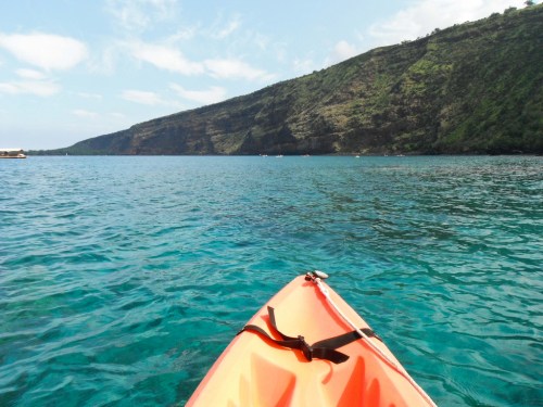 A vibrant orange kayak floats on clear blue water, with a lush green hillside in the background under a bright sky.
