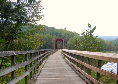 A wooden bridge leads to a red arch bridge surrounded by lush green trees and a cloudy sky.