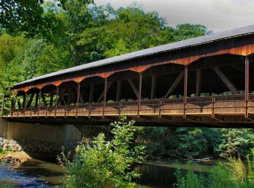 A wooden covered bridge spans a river, surrounded by lush greenery and trees.