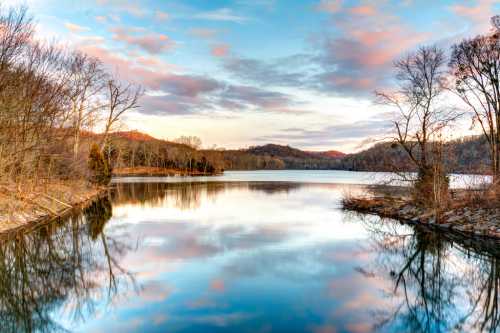 A serene lake reflects colorful clouds and trees, surrounded by hills under a tranquil sky at sunset.