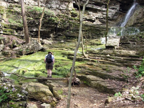 A hiker stands near a mossy rock formation and waterfall in a lush, green forest setting.