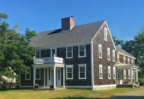 A large, two-story house with dark shingle siding, white trim, and a front porch, surrounded by greenery under a clear blue sky.