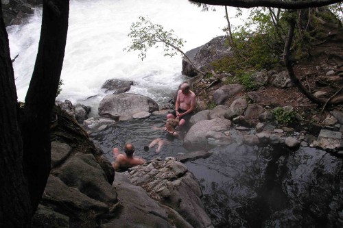 A group of people relax in a natural hot spring surrounded by rocks and trees, with a river flowing nearby.