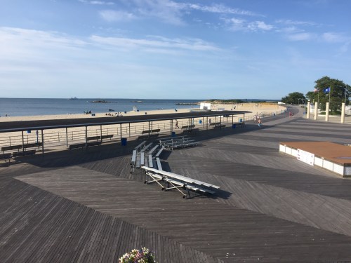 A sunny beach boardwalk with empty lounge chairs, a sandy shore, and calm ocean waves under a clear blue sky.