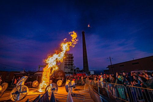 A crowd watches a large fire erupt from a structure at dusk, with industrial buildings and a smokestack in the background.