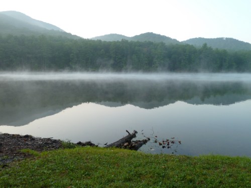 A serene lake surrounded by misty mountains, reflecting the landscape in calm waters. Green grass in the foreground.