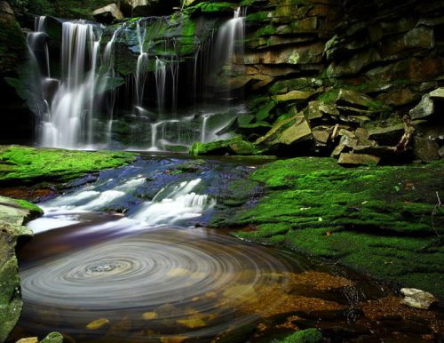 A serene waterfall cascades over moss-covered rocks into a tranquil pool, creating swirling patterns in the water.