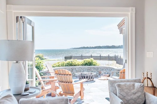 A view of a serene coastal scene through an open doorway, featuring wooden chairs and a sunny sky.