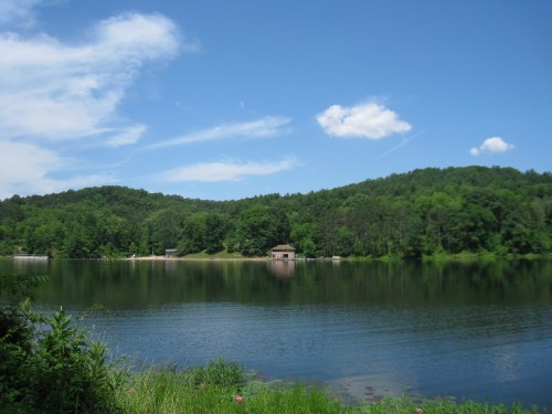 A serene lake surrounded by lush green hills under a clear blue sky, with a small building on the shore.